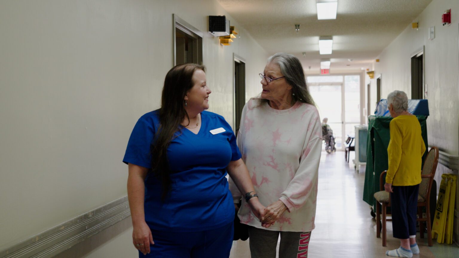 A woman smiles at a nurse as they walk down the hallway of a skilled nursing facility